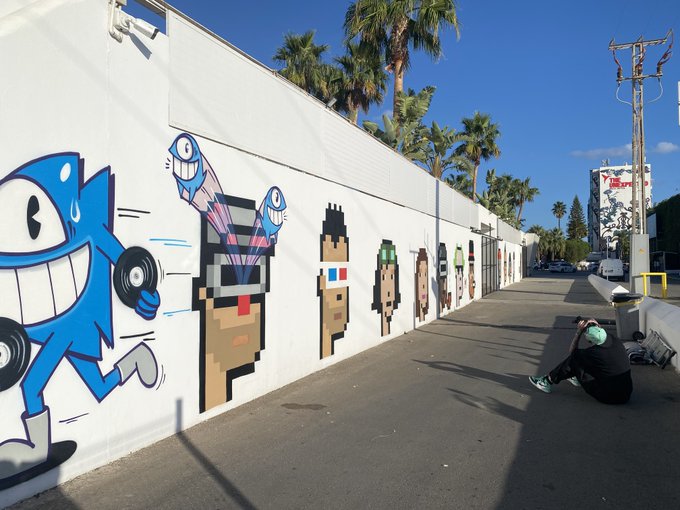A street-level photograph of a long mural painted on a white wall, featuring colorful cartoon characters, as a person sits on the sidewalk in the foreground near palm trees.