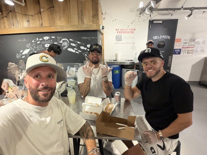 Three men pose at a counter inside "CULL & PISTOL" restaurant, with one making rock-on hand gestures and another flashing a fist, near a sign reading "WE VALUE YOUR FEEDBACK" and a QR code.