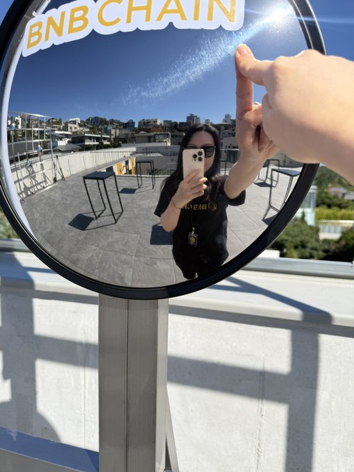 A mirror selfie reflection showing a person taking a photo with a smartphone in front of a round 'BNB CHAIN' sign on a rooftop terrace.