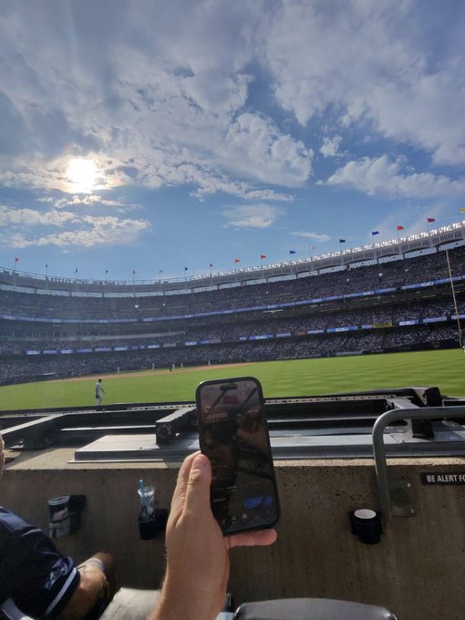 A first-person view from the stands of a packed baseball stadium under a sunny, cloudy sky, with a hand holding a smartphone in the foreground to capture the game.