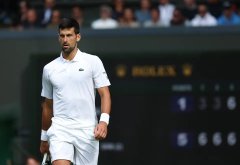 Tennis player Novak Djokovic walks on a grass court wearing a white outfit and headband, with a Rolex scoreboard visible in the background.