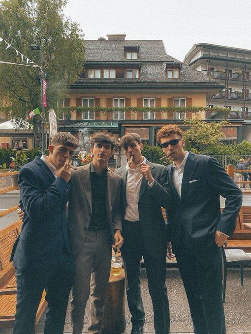 Four young men in suits pose together in an outdoor urban setting, with two smoking cigars.