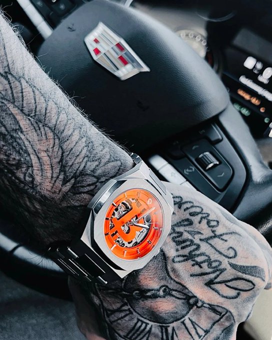 A close-up shows a heavily tattooed arm wearing a luxury skeleton watch with an orange dial, resting on the center console of a Cadillac vehicle.