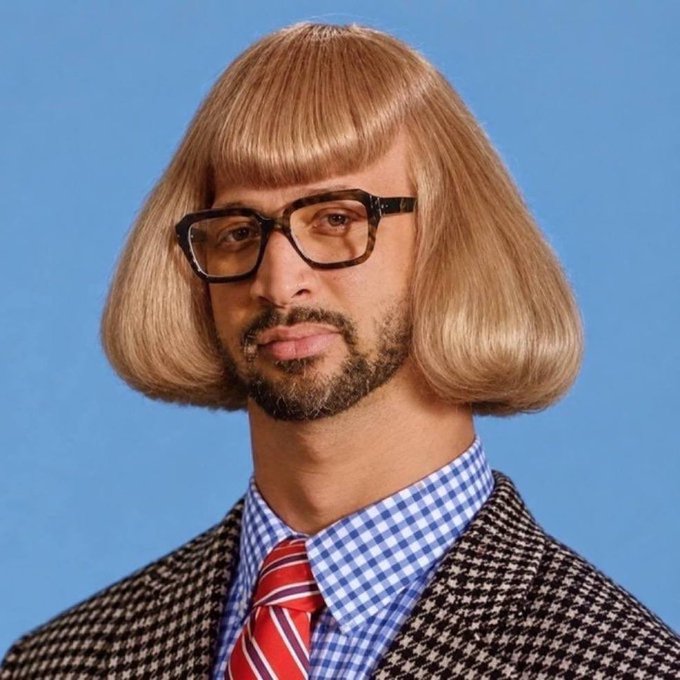 A man wears a comical blonde bob wig, thick-rimmed glasses, a checkered shirt, red tie, and houndstooth jacket against a blue background.