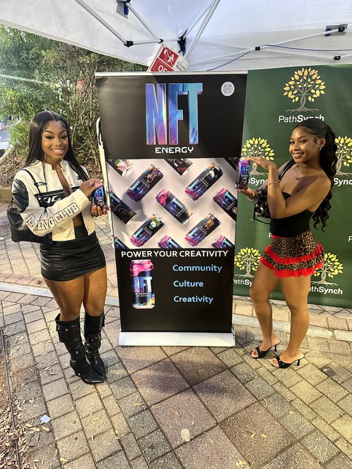 Two women pose outdoors beside an NFT Energy promotional display and a PathSynch backdrop, holding small colorful cans. Readable text includes “NFT ENERGY” and “POWER YOUR CREATIVITY,” with “Community,” “Culture,” and “Creativity” listed below.