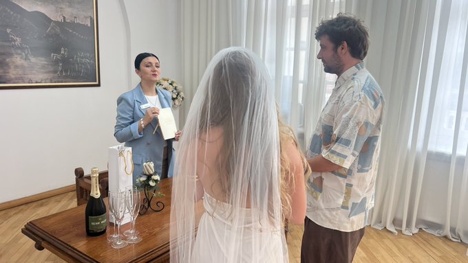 A bride in a white gown and veil stands with a man during a small indoor ceremony while an officiant in a light blue suit reads from a booklet. A table holds champagne, glasses, and a gift bag.