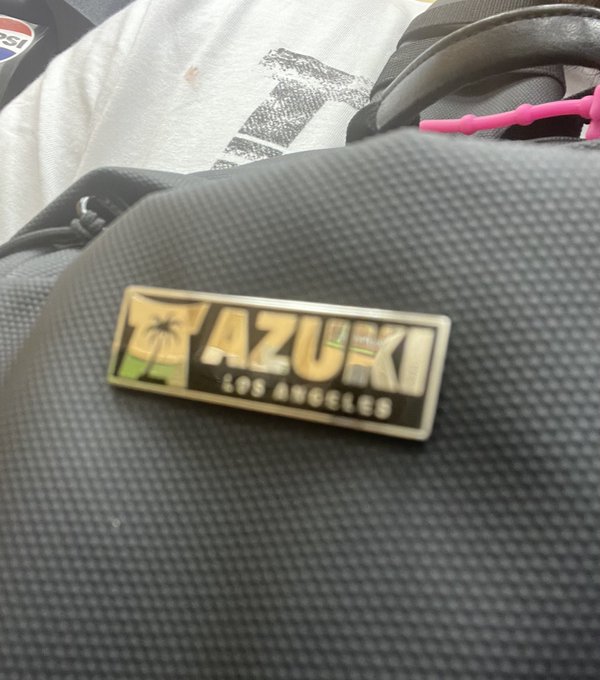 Close-up photo of a black textured bag with a rectangular gold and black enamel pin reading "AZUKI LOS ANGELES" featuring a palm tree design, with a white t-shirt and pink item visible in the background.