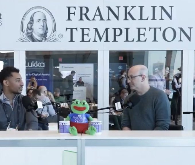Two men conducting an interview in front of a Franklin Templeton booth, with a Kermit the Frog plush toy wearing an 'Analytics for a Reason' shirt sitting on the counter between them.