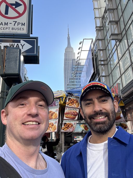 Two smiling friends take a selfie on a sunny Midtown Manhattan street, with the Empire State Building towering in the background alongside a typical NYC hot dog cart advertising items like "Jumbo Hot Dog" and "Chicken & Fries." Street signs indicating "7 AM-7 PM EXCEPT BUSES" and a one-way arrow frame the iconic urban scene.