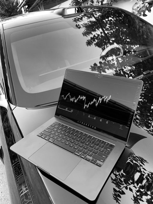A black and white photograph shows a MacBook displaying a financial chart resting on the hood of a sleek car, with tree reflections visible on the vehicle's surface.