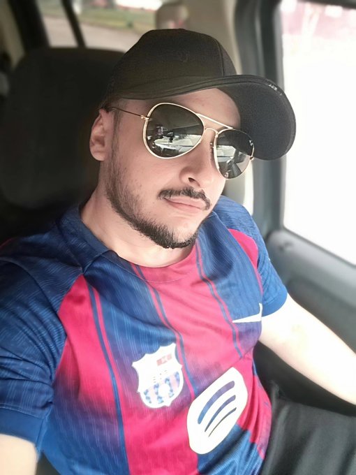 A selfie of a man wearing a black cap, sunglasses, and a blue and red striped football jersey, sitting inside a vehicle.