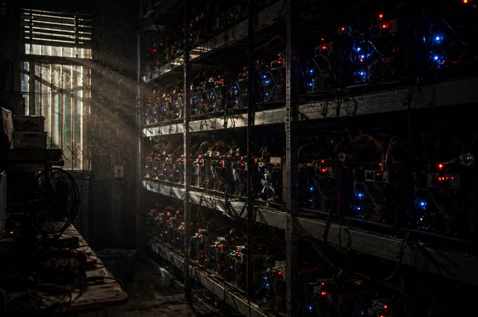 Dusty sunbeams filter through a grim window onto towering shelves of GPU mining rigs, their status lights glowing red and blue in a dim, industrial room. The image visually echoes the post's point: each glowing rig represents the massive, tangible electrical cost that physically secures a Proof-of-Work network, making a 51% attack prohibitively resource-draining.