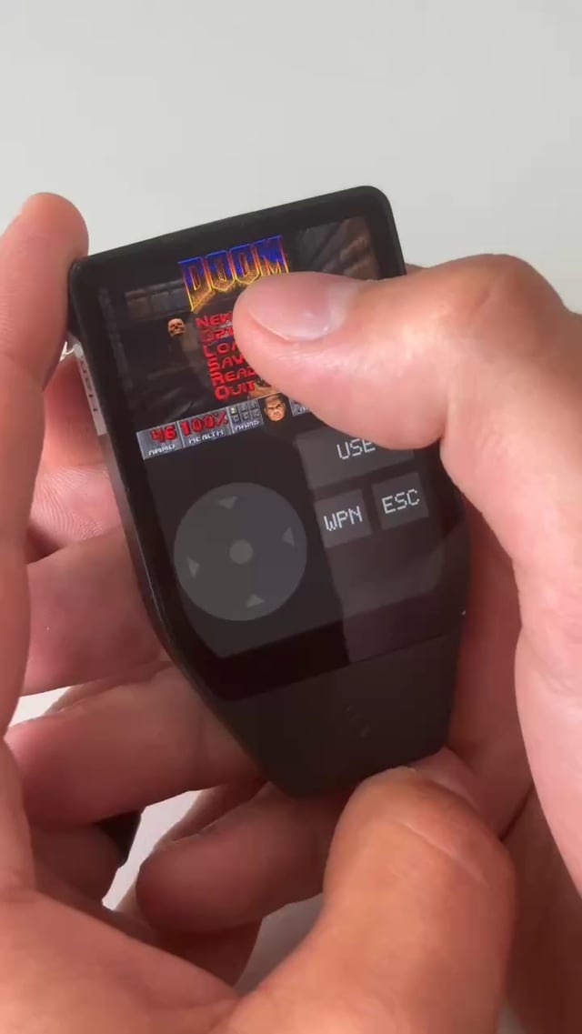 A hand holds a Trezor Safe 7 hardware wallet displaying the classic DOOM game on its small screen, with on-screen touch controls including a directional pad and buttons labeled "USE," "WPN," and "ESC," alongside the game's iconic red "DOOM" logo and 100% health indicator.