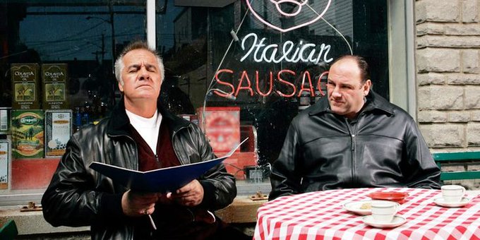 Two men in black leather jackets sit at a table outside an Italian sausage shop; one man holds a blue book or folder.