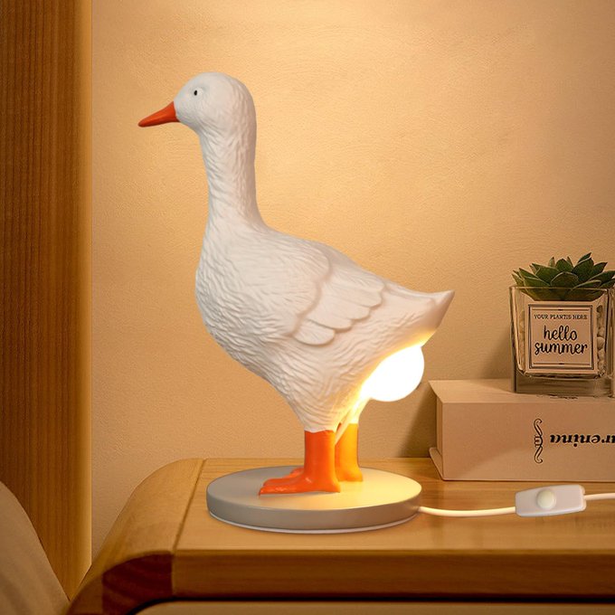 A white goose-shaped table lamp with orange beak and feet sits on a wooden nightstand next to a small succulent labeled 'hello summer' and a book.