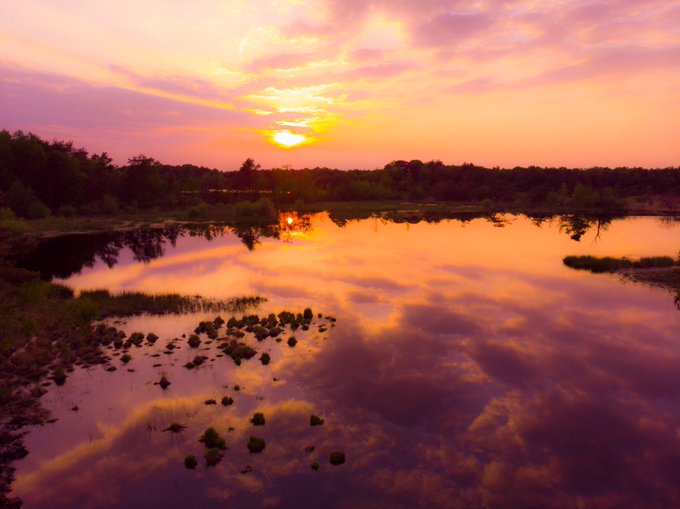 A vibrant sunset reflecting off a calm river, with orange and pink clouds mirrored in the water alongside silhouetted trees on the riverbank.