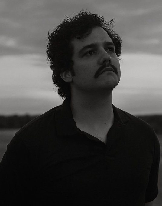 A black and white portrait of a man with curly dark hair and a mustache wearing a polo shirt, gazing off to the side against a cloudy sky background.
