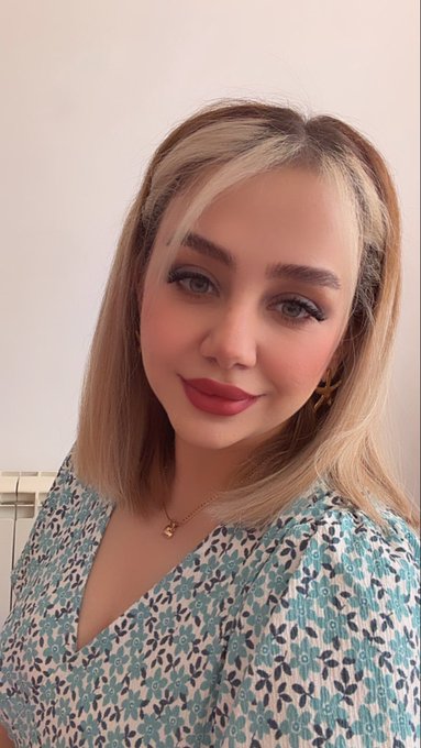 A young woman with long blonde hair poses for a selfie, wearing a floral-patterned top with teal and white tones. The image serves as a profile photo accompanying a post about finding an owner for an NFT artwork titled "The Pulse of Nature" priced at 80 $XTZ.