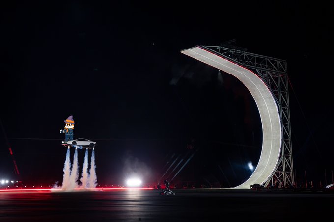 A nighttime stunt shows a car being launched into the air by jet propulsion toward a massive curved ramp, with flames and smoke trailing behind it on the ground.