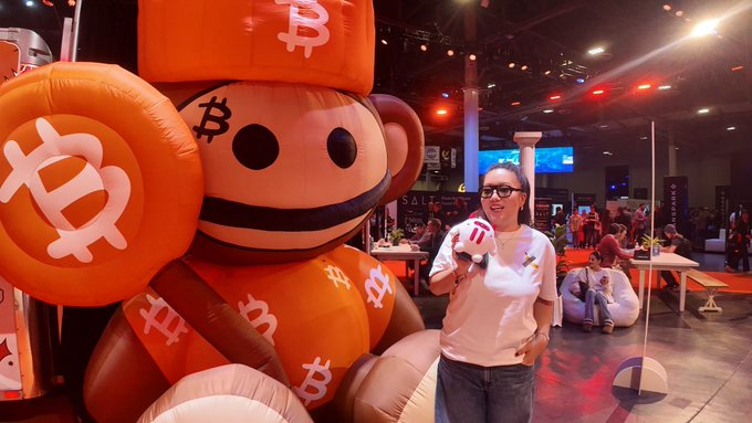 A woman poses beside a large inflatable Bitcoin mascot at a cryptocurrency conference, with "SALT" visible on signage in the background. The image's Bitcoin branding ironically contrasts with the post's announcement about TRON DAO's new on-chain green energy certification governance for ESG-compliant blockchain development, highlighting the industry's broader pivot toward sustainability.