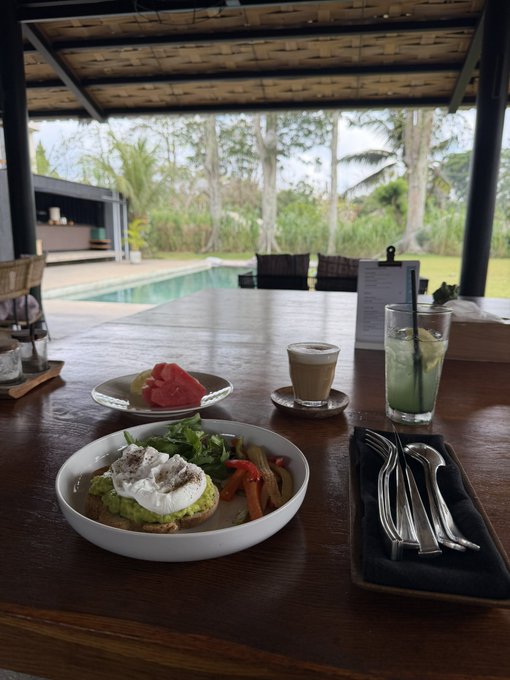 A breakfast spread including avocado toast with poached eggs, fresh fruit, a latte, and a green juice, set on a wooden table overlooking a pool and lush tropical landscaping.