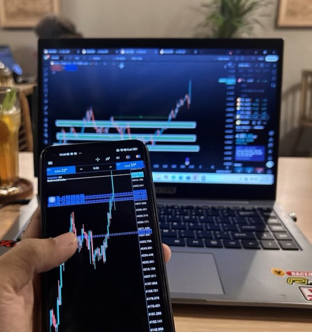 A person holds a smartphone displaying a financial trading chart with values around 4204.32 and 6.86, positioned in front of a laptop showing similar trading software.