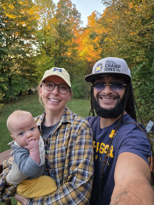 A selfie of a couple wearing University of Michigan apparel (a woman with a yellow 'M' cap and plaid shirt holding a baby, and a man with a 'CHAMPIONS' cap and Michigan shirt) outdoors in autumn.