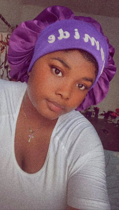 A selfie of a woman wearing a purple satin bonnet with white lettering and a light blue top with a cross necklace.