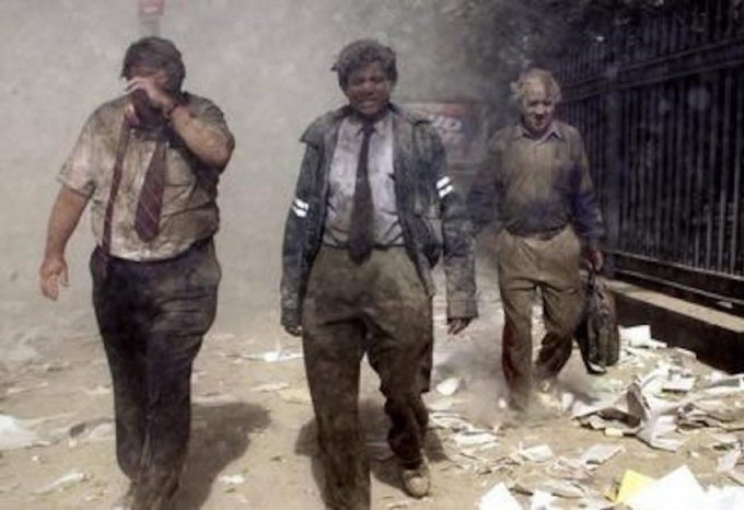 Three men in dirty business attire walk through a dusty, debris-filled street amid smoke and scattered papers, evoking a disaster scene.