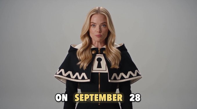 A blonde woman in a dark hooded outfit with a keyhole symbol on the chest, with text reading 'ON SEPTEMBER 28' overlaid on the image.