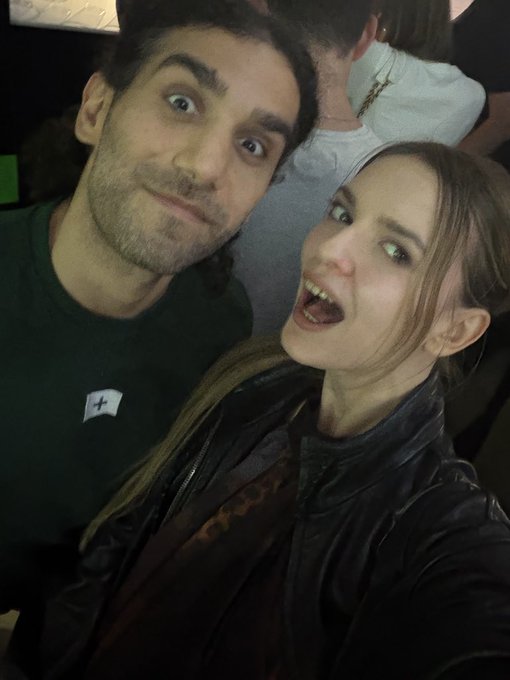 A man and a woman take a selfie together; the man has wide eyes and a slight smile, while the woman smiles widely with her mouth open.