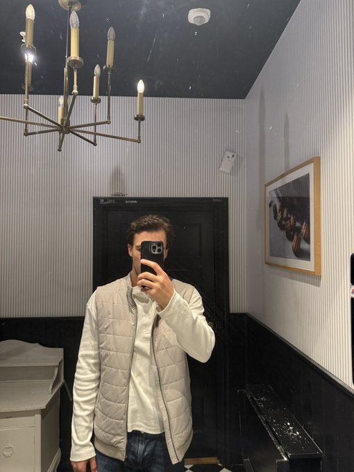 A person takes a mirror selfie while wearing a light-colored quilted vest over a sweater, standing in a room with a chandelier, striped walls, and framed artwork.