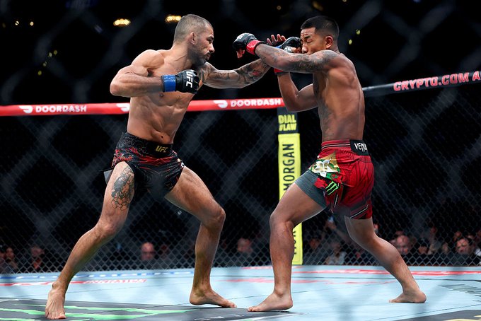 Two UFC fighters engage in a fierce stand-up exchange inside the octagon, with one in black shorts landing a punch against an opponent in red and black Venum shorts, as "DOORDASH," "CRYPTO.COM," and "UFC" branding are visible on the canvas and cage. The intense striking action illustrates the "salvaje primer round" (savage first round) caption accompanying the #CryptoCom #UFC331 post.