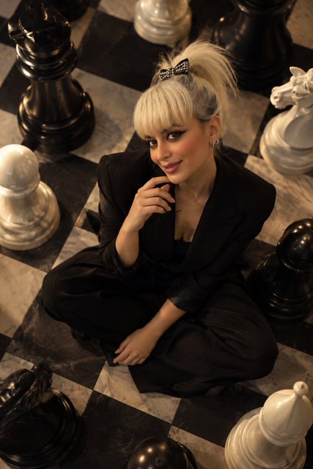 A woman with blonde hair in a high ponytail with a checkered bow sits cross-legged on a marble chessboard surrounded by giant black and white chess pieces, wearing a black suit.