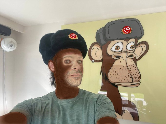 A man wearing a furry black Bored Ape Yacht Club (BAYC) hat poses next to a cartoon monkey mural of his NFT ape, showcasing his first BAYC purchase to the community.