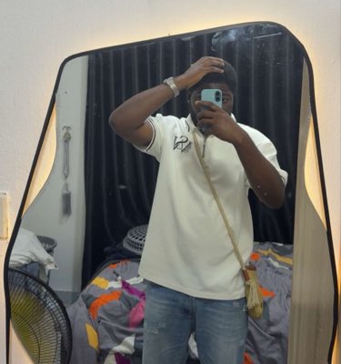 A man wearing a white t-shirt with an "AM" logo, jeans, and a crossbody bag takes a mirror selfie with his phone in a bedroom featuring dark curtains and a colorful blanket. The casual, anonymous nature of the mirror shot fits the post's theme of promoting a secondary "alt" social media account to engage in provocative or disruptive conversations.