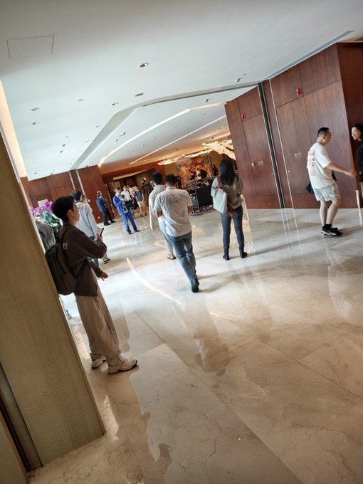 A photograph taken inside a hotel lobby or similar building, showing several people standing and walking on a polished marble floor with wood-paneled walls.