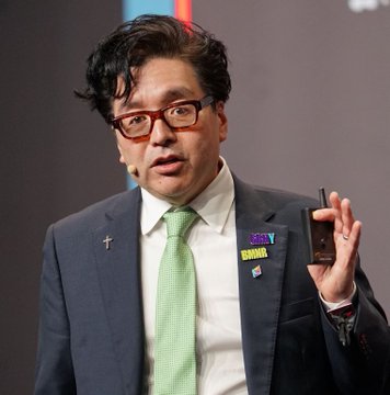 A man with dark hair and glasses, wearing a grey suit with a green tie and a '2025' lapel pin, holds a small black device while speaking.