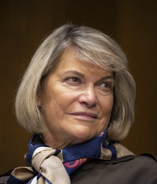 The image shows a close-up of Senator Cynthia Lummis, identified by the accompanying post, who is pictured wearing a tan jacket and a blue patterned scarf while looking off to the side with a slight smile. The caption relates to her recent criticism of banks opposing the Clarity Act's stablecoin regulations.