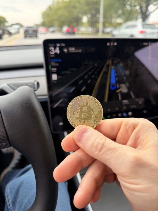 A hand holding a Bitcoin (BTC) gold coin in front of a Tesla touchscreen display showing navigation while driving on a road with other vehicles visible.