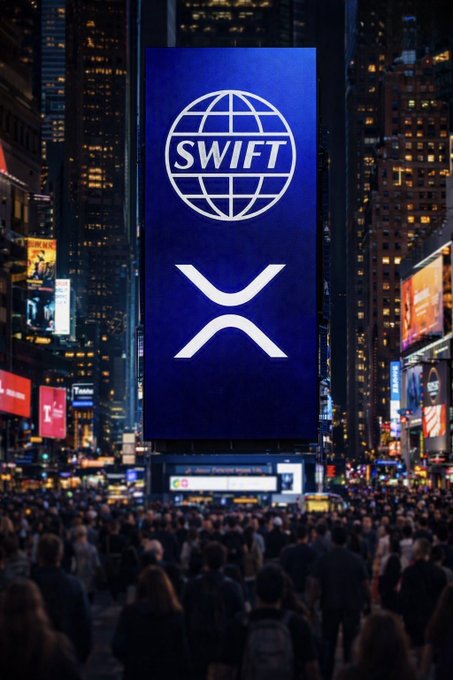 A large digital billboard in a crowded urban area at night displays the SWIFT logo and the XRP cryptocurrency symbol against a dark blue background.