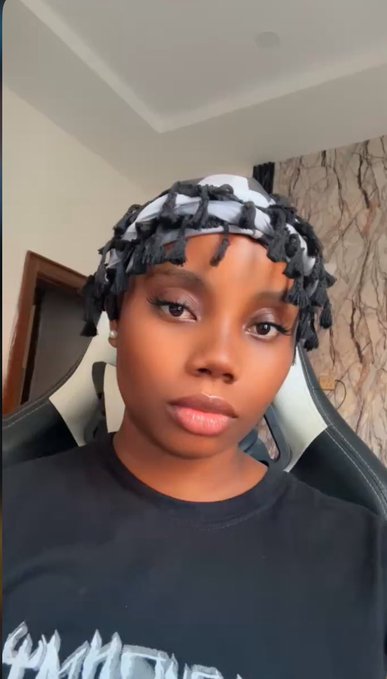 A selfie of a young woman wearing a black t-shirt and a patterned head wrap with black tassels, sitting in a gaming chair indoors.