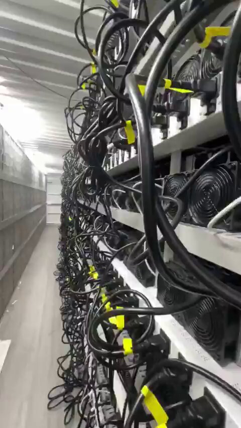 The image shows an indoor space filled with numerous black cables, likely used for electrical purposes or as part of a computer network. The cables are connected to various electronic devices such as computers and other equipment. In the background, there is a white wall that provides contrast to the dark cables. A yellow label can be seen on one of the cables, possibly indicating its purpose or location within the space.