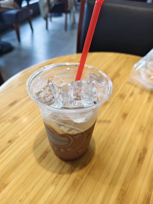 An iced coffee in a clear plastic cup with a red straw sits on a wooden table, with the 'NO' and 'COFFEE' text visible on the cup's logo.