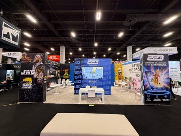The image shows a trade show booth for the Licensing Expo at the convention center. The booth features a large screen TV displaying an advertisement for "@RIPNDIP". There are two chairs placed near the booth, one of which has a person sitting on it. A couch is also present in the scene. The booth appears to be well-organized and visually appealing, showcasing various products or services related to IPs and licensing.