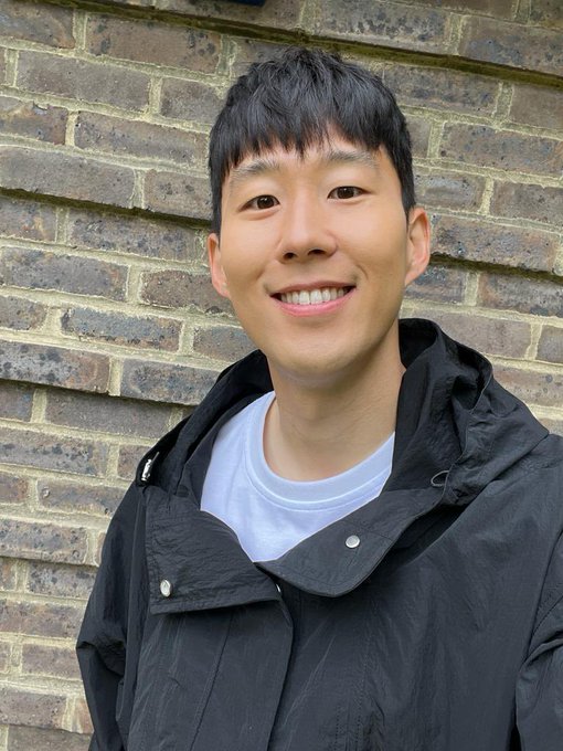 A portrait photograph shows a smiling young Asian man wearing a white t-shirt under a black hooded jacket, standing in front of a tan brick wall.