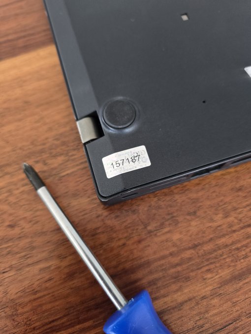 A close-up of the back of a black laptop on a wooden surface, showing a serial number sticker (157187...SN04...) near the hinge, with a blue-handled screwdriver placed beside it.