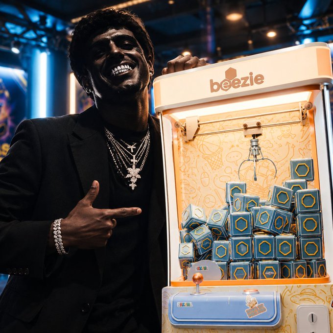 A man wearing a dark suit and multiple cross necklaces points to a 'beezie' branded claw machine filled with blue hexagonal boxes.
