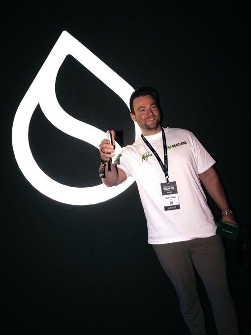 A cheerful man wearing a lanyard gives a thumbs-up next to the illuminated Sui Network logo at the Sui Network Basecamp booth during Token2049, posing in front of the brand's large illuminated water drop signage.
