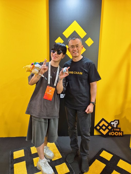 Two attendees pose together in front of a Binance-branded yellow backdrop featuring the platform's diamond logo, with one wearing a "BNB CHAIN" t-shirt and both flashing peace signs alongside plush toys. The image captures the friendly, optimistic atmosphere of the "GM Legends" crypto community greeting the day with smiles and good vibes.